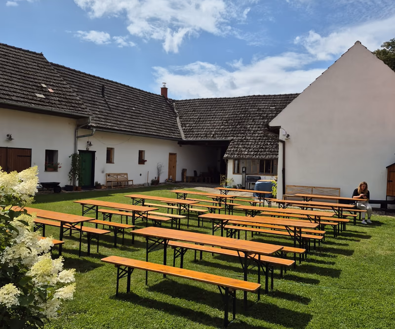 Dvůr Statku Žíšov s dřevěnými stoly a lavicemi na trávníku mezi bílými budovami